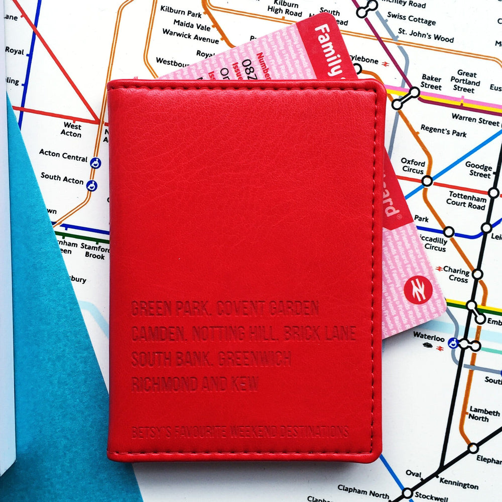 A red personalised vegan leather card holder is lying closed on top of a London Underground tube map. Poking out the side is a family railcard. The front of the travelcard cover is engraved with 5 lines of personalised text detailing the travellers favourite London tube stops. The last line reads Betsys favourite weekend destinations.