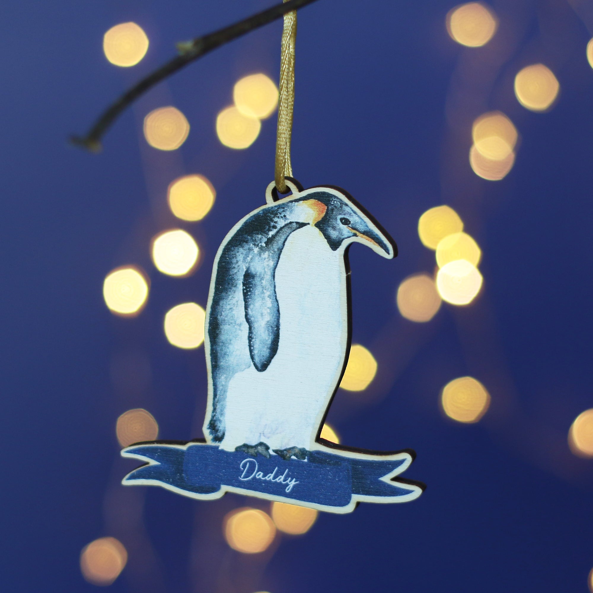 A wooden penguin decoration hangs from a branch. It is a watercolour of an Emperor penguin and has the name Daddy at the bottom. In the background there's a blue wall and twinkly Christmas lights