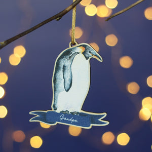 A wooden penguin decoration hangs from a branch. It is a watercolour of an Emperor penguin and has the name Grandpa at the bottom. In the background there's a blue wall and twinkly Christmas lights