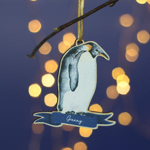 A wooden penguin decoration hangs from a branch. It is a watercolour of an Emperor penguin and has the name Granny at the bottom. In the background there's a blue wall and twinkly Christmas lights