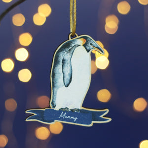 A wooden penguin decoration hangs from a branch. It is a watercolour of an Emperor penguin and has the name Mummy at the bottom. In the background there's a blue wall and twinkly Christmas lights
