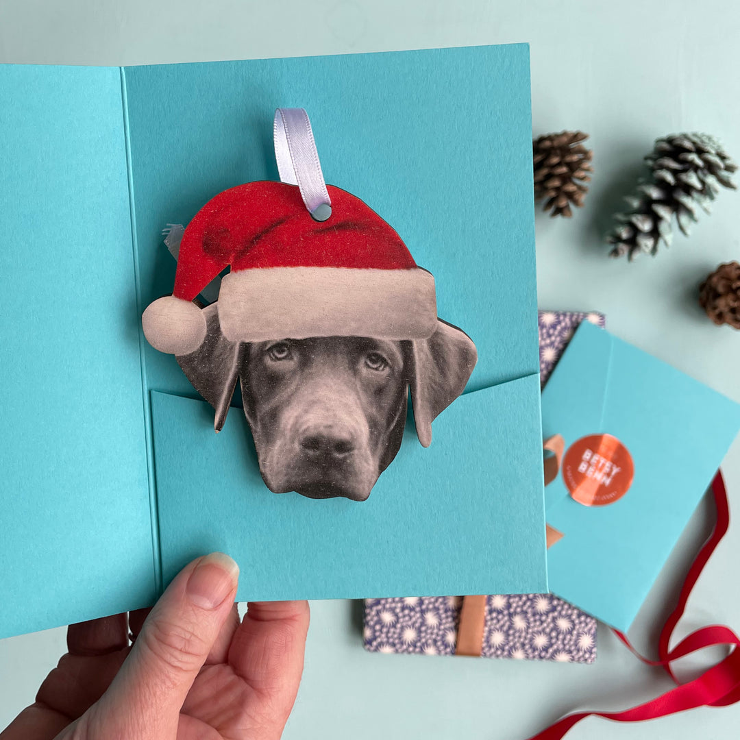 Christmas decoration of a dog wearing a Santa hat with a white satin ribbon for hanging, being packed in a gift sleeve. 