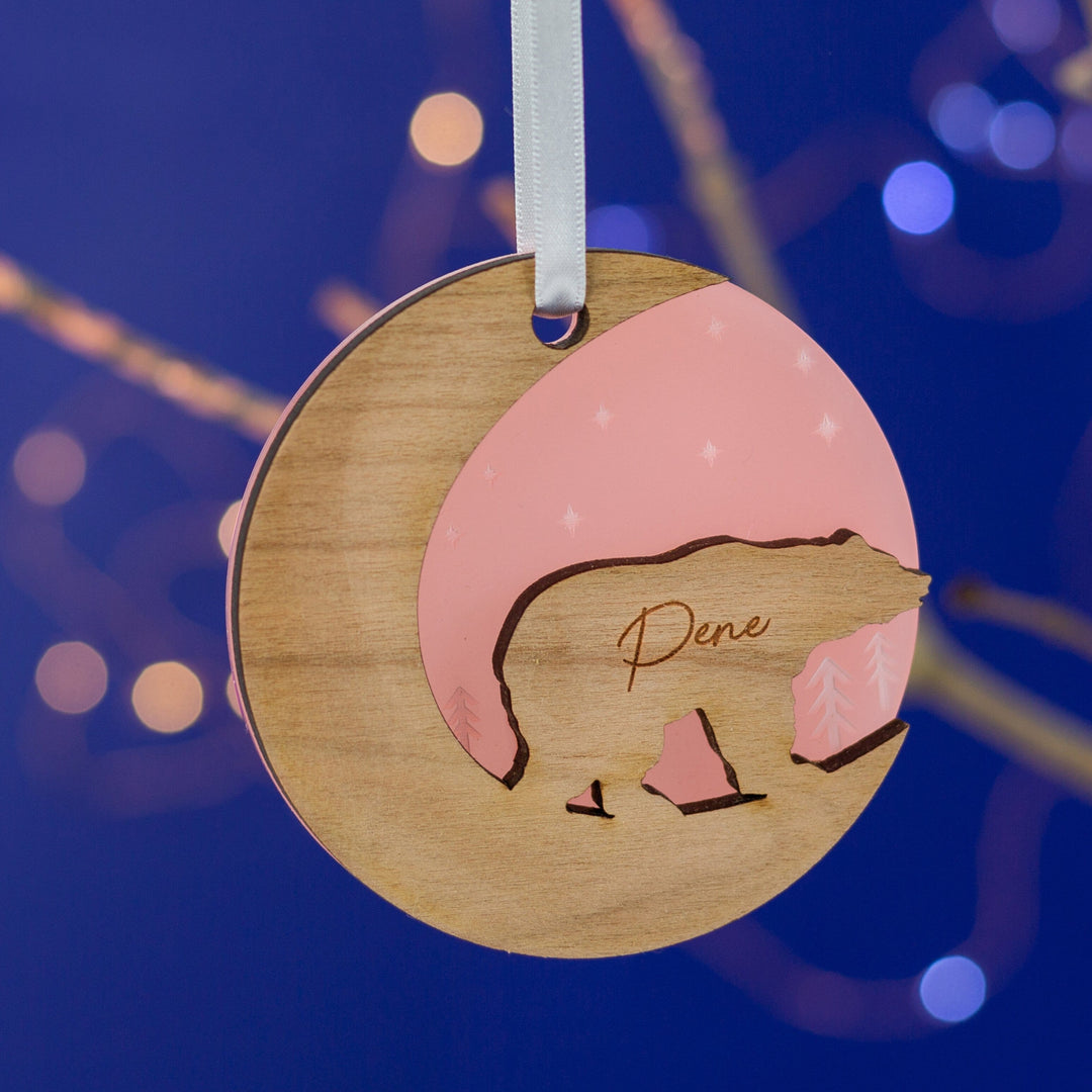 A round wooden Christmas ornament hangs against a dark blue background with bokeh lights. The ornament features a bear silhouette and is engraved with the name "Dene." It has a crescent moon cutout and a pink inset with stars.