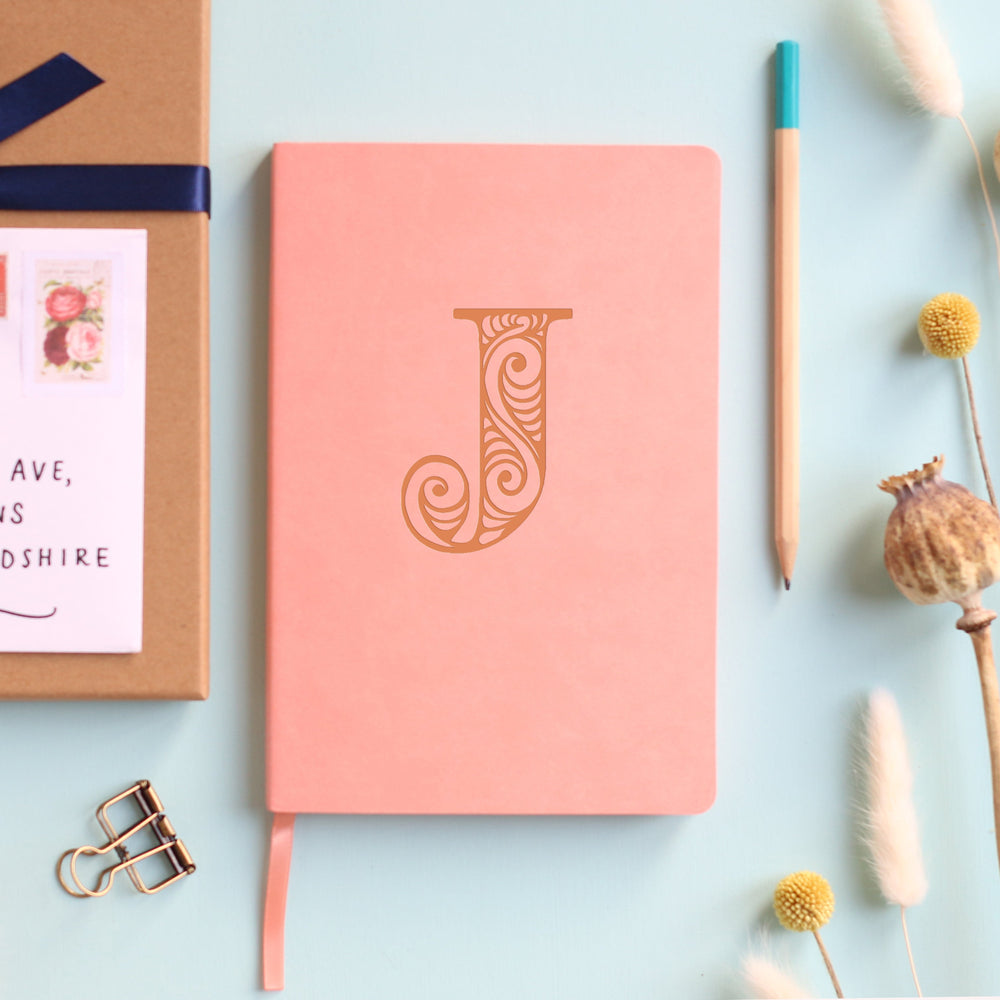 A top down image of a pink vegan leather notebook resting on a pale aqua table. The front of the notebook is personalised with the letter J engraved in a large decorative font. Surrounding the book are various stationary items and dried flowers.
