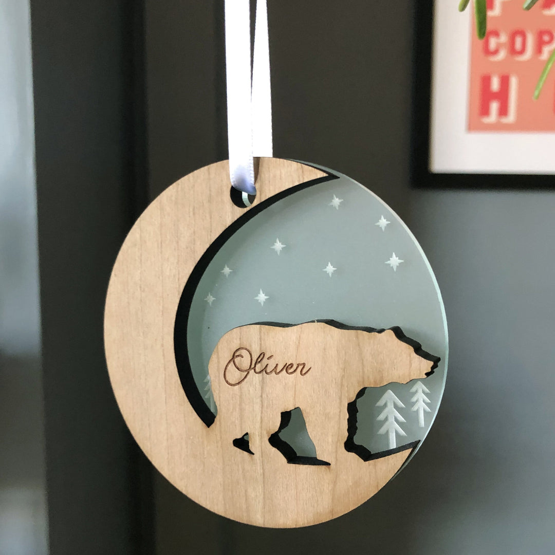 A round wooden Christmas ornament hangs against a blurred background with a framed picture. The ornament features a bear silhouette and is engraved with the name "Oliver." It has a crescent moon cutout and a light inset with stars.