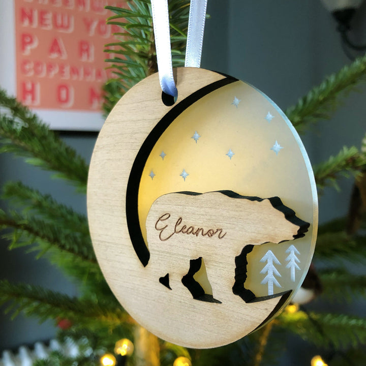 A round wooden Christmas ornament hangs on a pine branch with a blurred background. The ornament features a bear silhouette and is engraved with the name "Eleanor." It has a crescent moon cutout and a light inset with stars. A postcard with city names is partially visible in the background.