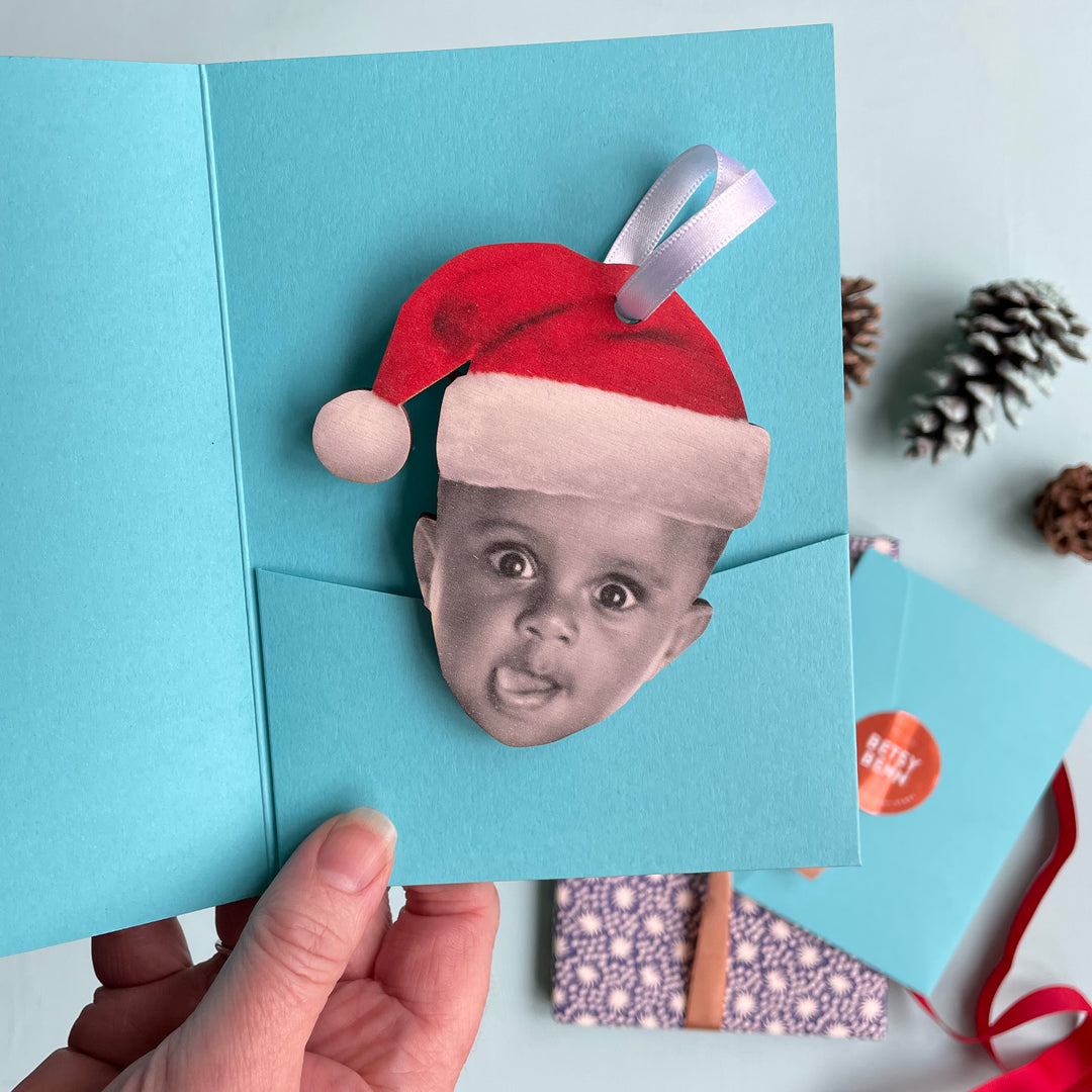 Wooden Christmas bauble featuring a toddler character in a Santa hat, festive keepsake with blue gift sleeve in the background.