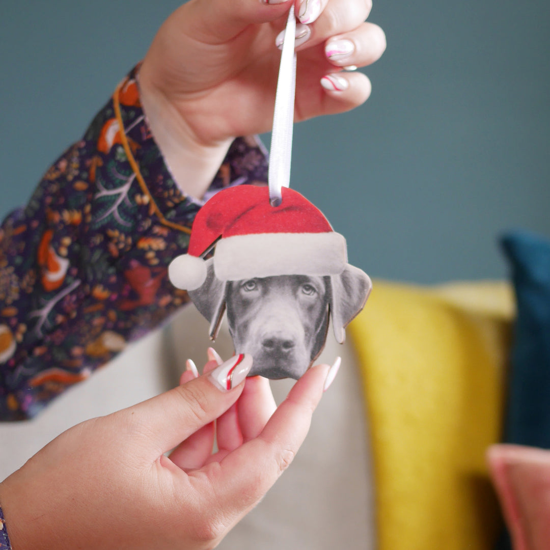Wooden custom Christmas decoration of a dog's head wearing a Santa hat, attached to a satin white ribbon for hanging, held by a hand, making it a perfect gift for dog lovers. 
