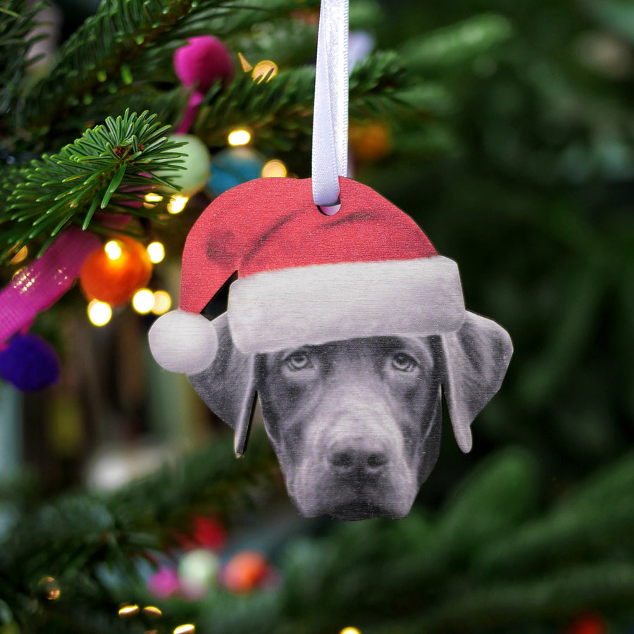 A wooden Christmas tree decoration featuring a black and white image of a dog wearing a Santa hat, with a white satin ribbon for hanging.