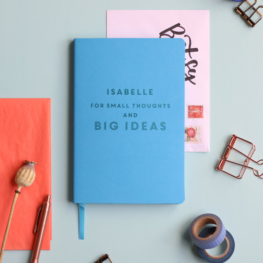 A top down image of a blue vegan leather notebook resting on a pale aqua table. The front of the notebook features the phrase "For small thoughts and big ideas". It is personalised with the name Isabelle engraved above the text. Surrounding the book are various stationary items and dried flowers.