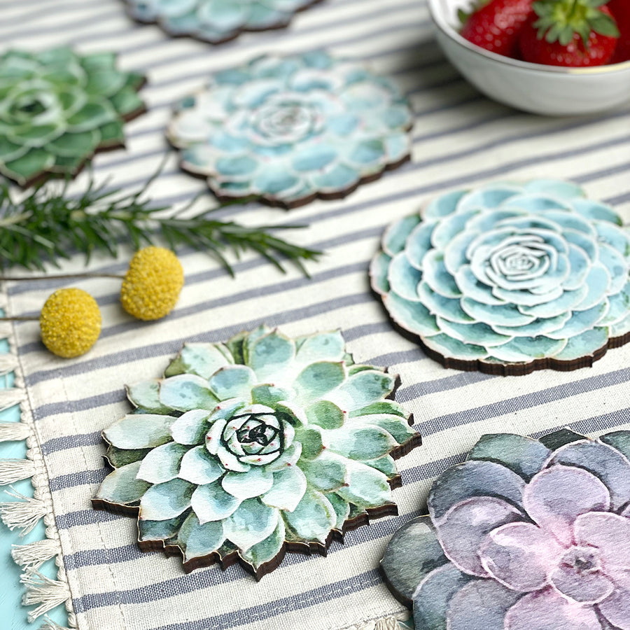 Succulent-shaped wooden coasters on a striped fabric with decorative elements, including greenery, yellow flowers, and a bowl of strawberries.