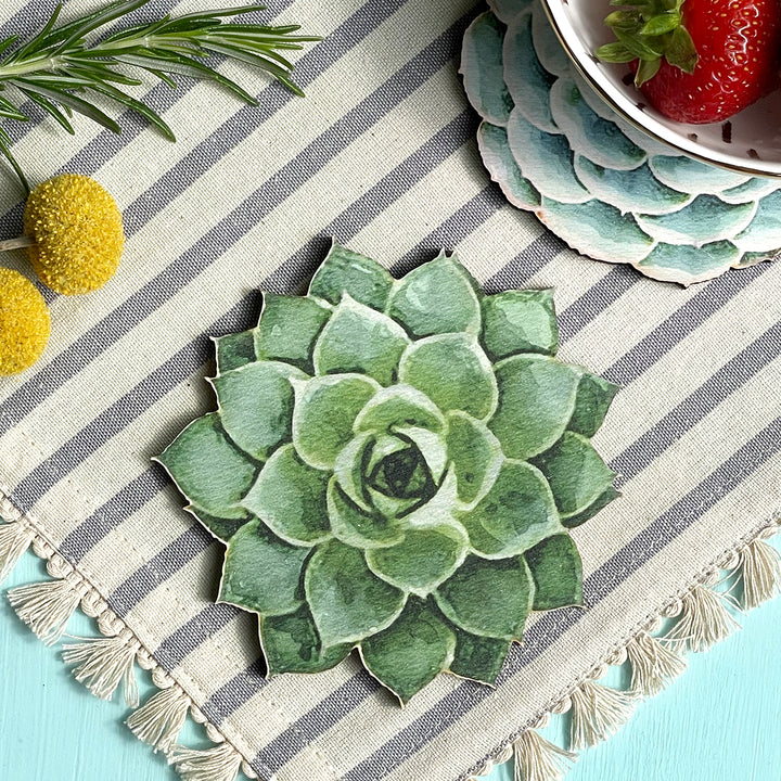 Close-up of a succulent-shaped wooden coaster on a striped fabric with decorative elements, including yellow flowers, rosemary, and a bowl of strawberries.