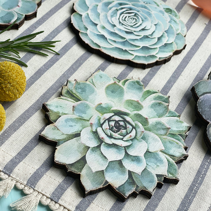 Wooden coasters shaped like succulents with a watercolor-style design on a striped fabric, decorated with rosemary and yellow billy button flowers.