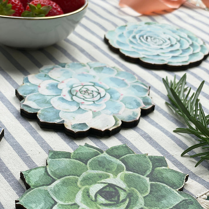 Succulent-shaped wooden coasters with watercolor-style designs on a striped tablecloth, accompanied by a bowl of strawberries and rosemary sprigs.