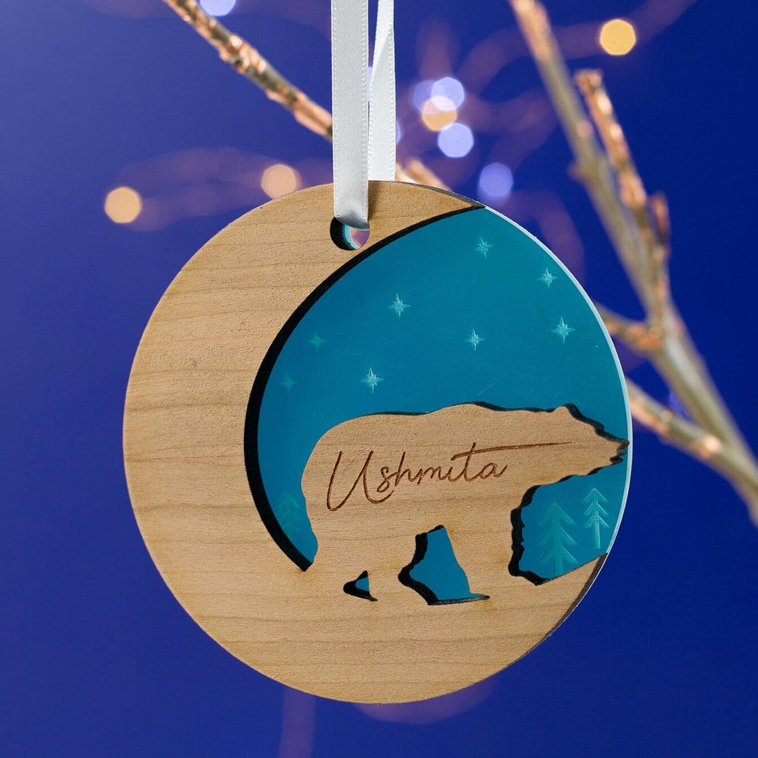 A round wooden Christmas ornament hangs on a branch against a dark blue background with bokeh lights. The ornament features a bear silhouette and is engraved with the name "Ushmita." It has a crescent moon cutout and a teal inset with stars.