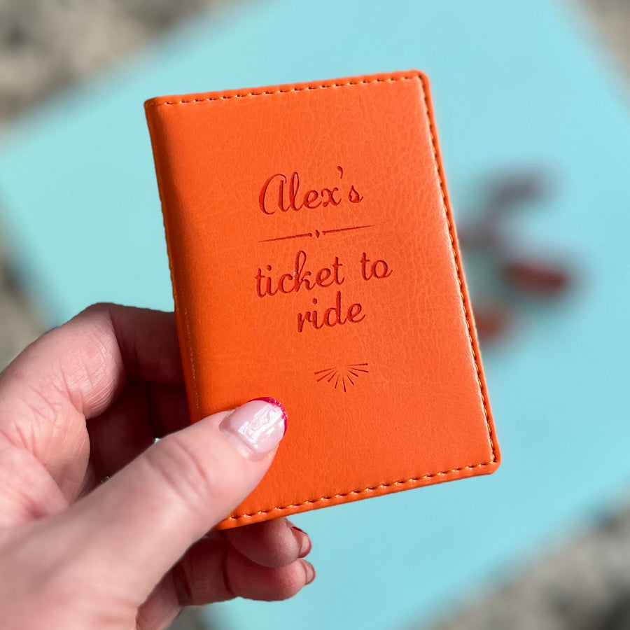 A hand holds a small orange leather card holder with the text "Alex's ticket to ride" engraved on the front. The holder has stitched edges and a small design at the bottom.