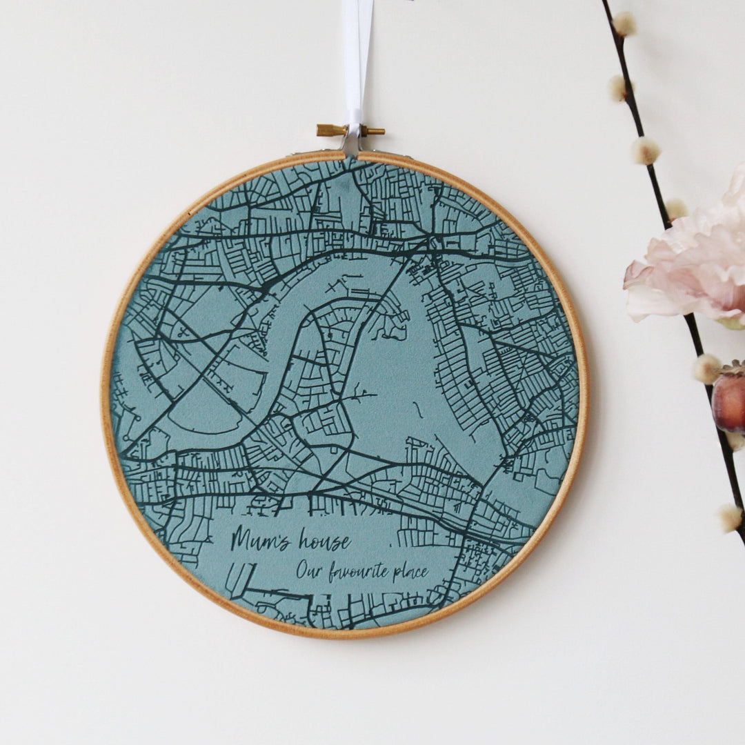 A round embroidery hoop with a teal fabric map of a city inside. The map includes roads, rivers, and the text "Mum's house. Our favourite place."