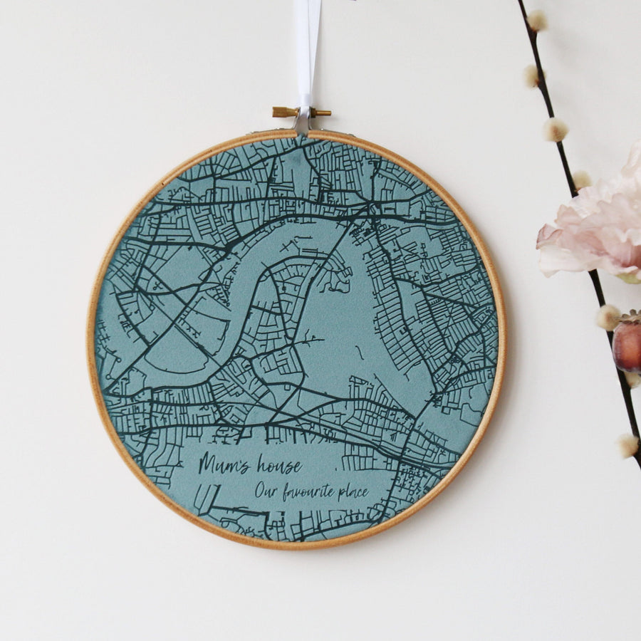 A round embroidery hoop with a teal fabric map of a city inside. The map includes roads, rivers, and the text "Mum's house. Our favourite place."
