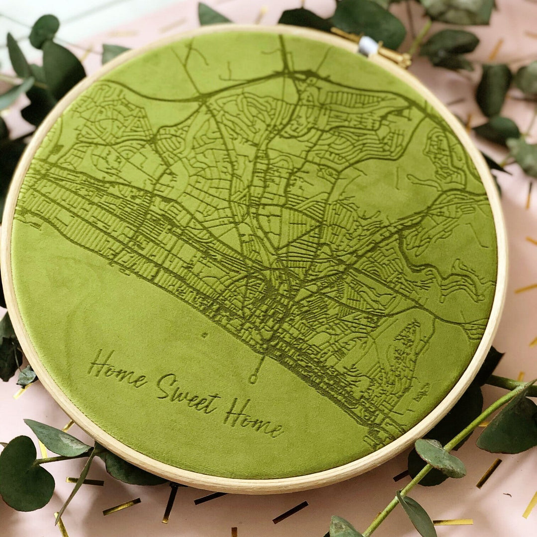 A circular embroidery hoop with a green velvet map of a city embroidered on it. The text "Home Sweet Home" is embroidered at the bottom. The hoop is surrounded by green leaves.