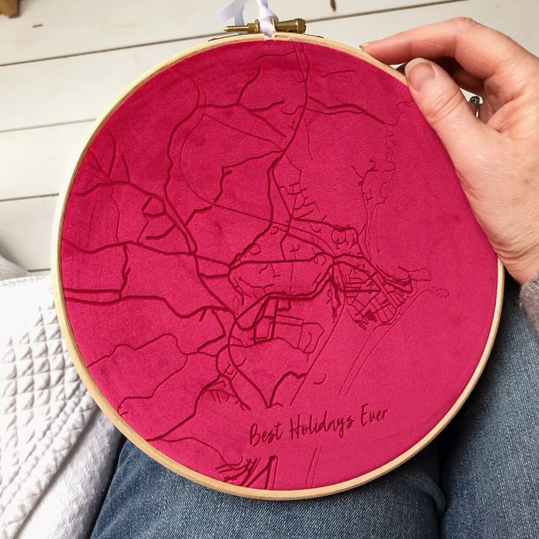 A hand holding a red velvet embroidery hoop with a map embroidered on it. The text "Best Holidays Ever" is embroidered at the bottom.