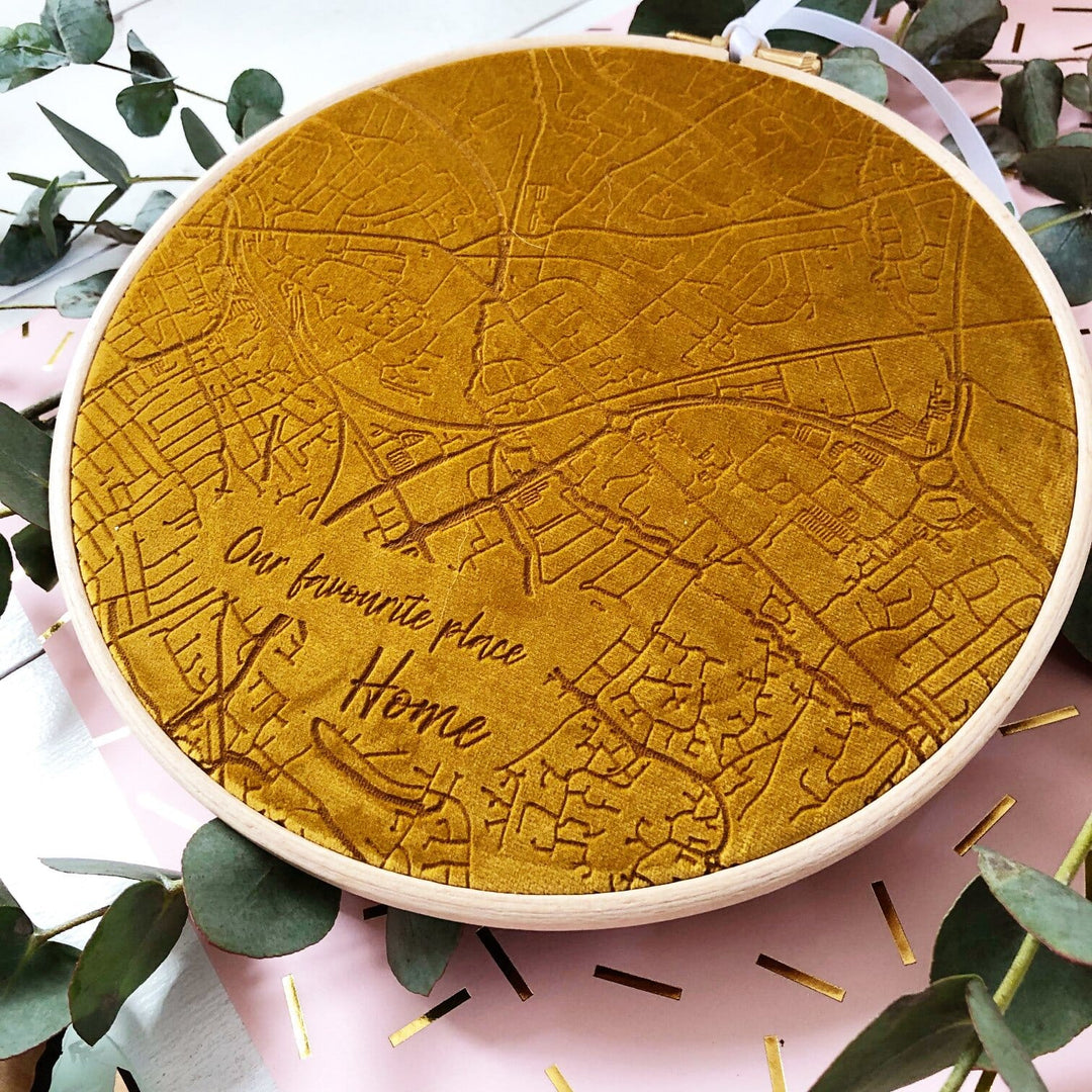A circular embroidery hoop with a mustard yellow velvet map of a city embroidered on it. The text "Our favorite place. Home" is embroidered at the bottom. The hoop is surrounded by green leaves.