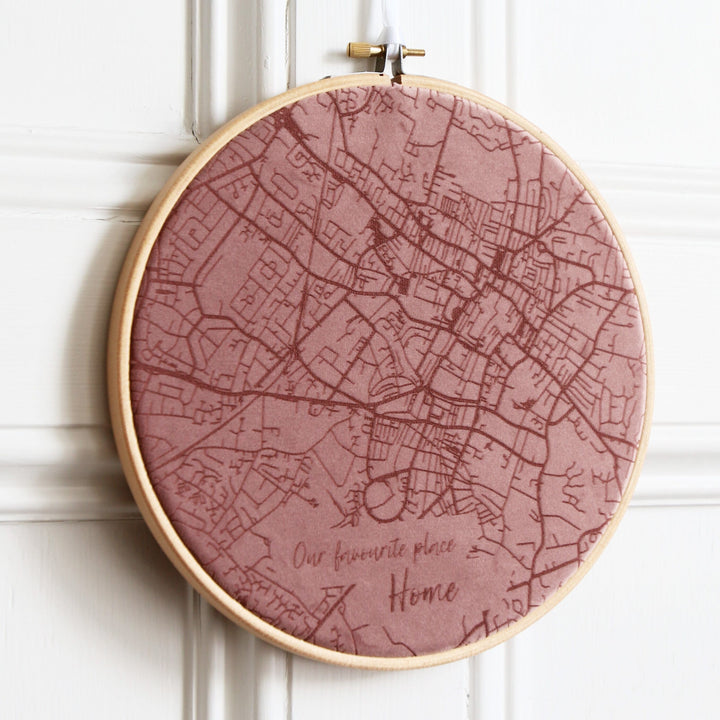 A round embroidery hoop with a pink velvet map of a city embroidered on it. The text "Our favourite place. Home" is embroidered at the bottom. The hoop is hanging on a white wall.