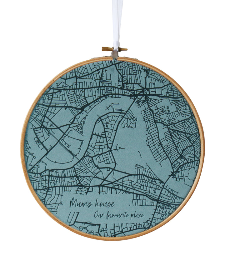 A round embroidery hoop with a teal fabric map of a city inside. The map includes roads, rivers, and the text "Mum's house. Our favourite place."