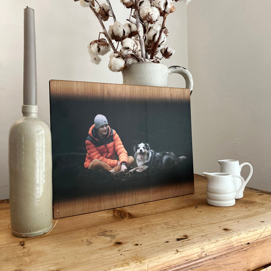 A wooden photo print of a person in an orange jacket sitting with a dog, displayed on a rustic table with decorative items.