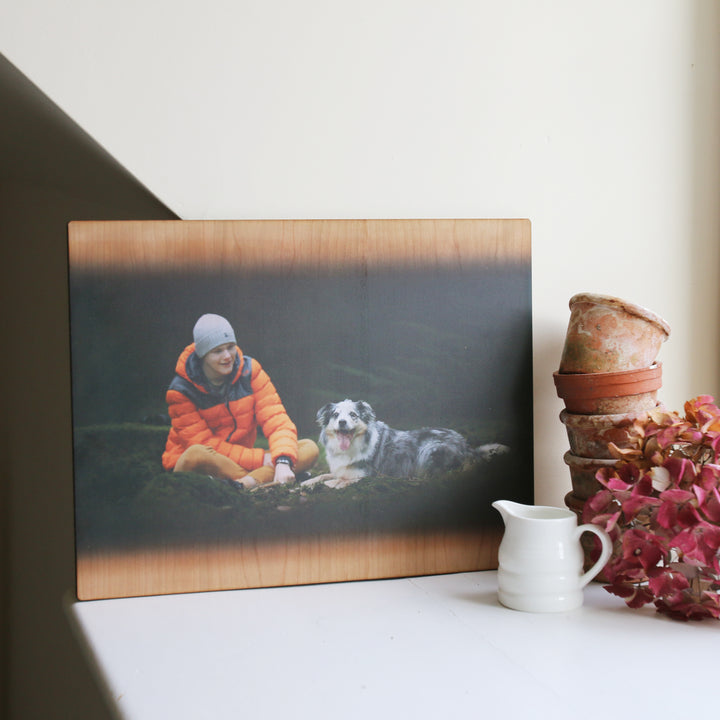 A wooden photo print of a person in an orange jacket sitting with a dog, displayed on a rustic table with decorative items.